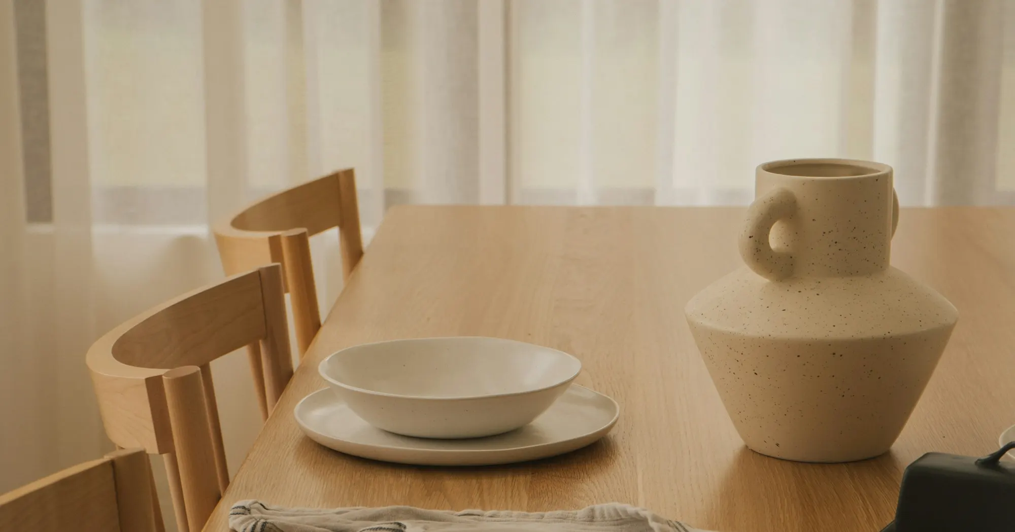 A hand-thrown speckled stoneware vessel and a white bowl on a plate, set on a timber table in front of sheer linen curtains.
