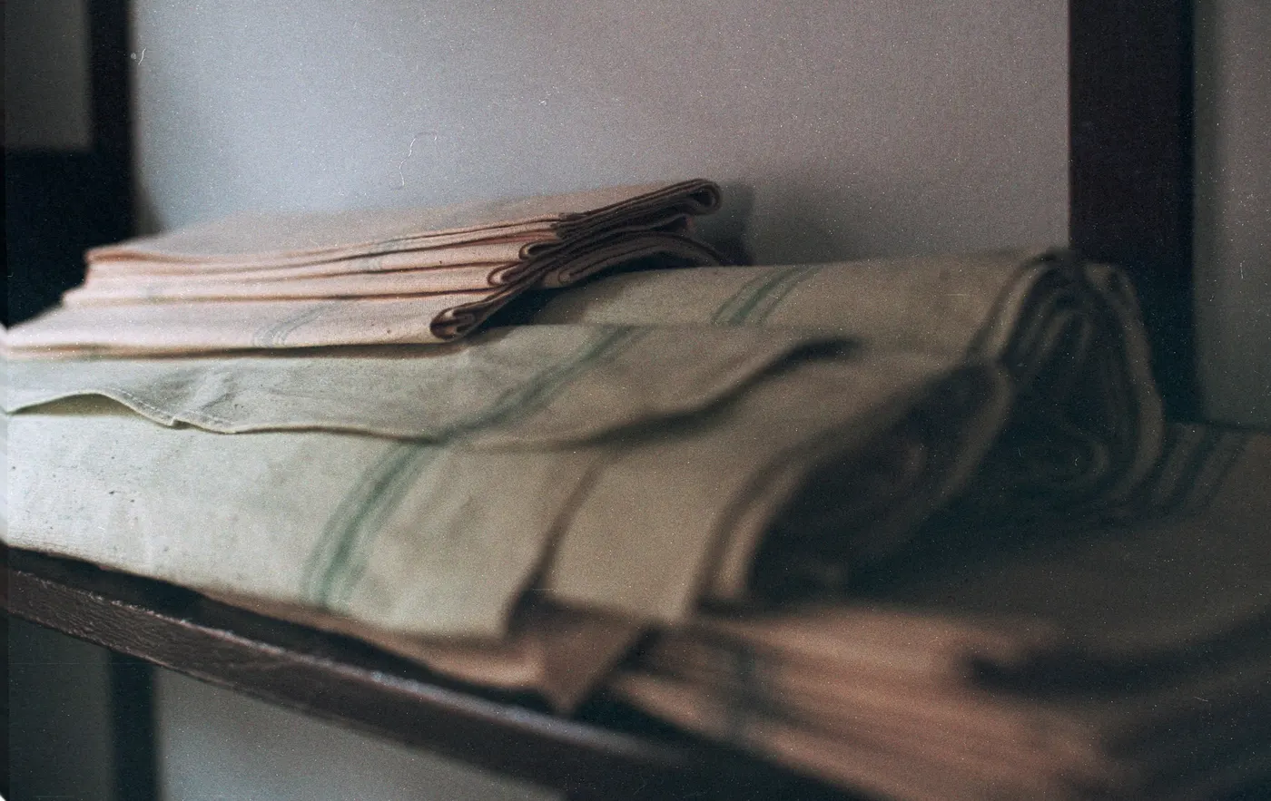 Folded linen cloths in flax and blush tones stacked on a dark timber shelf.