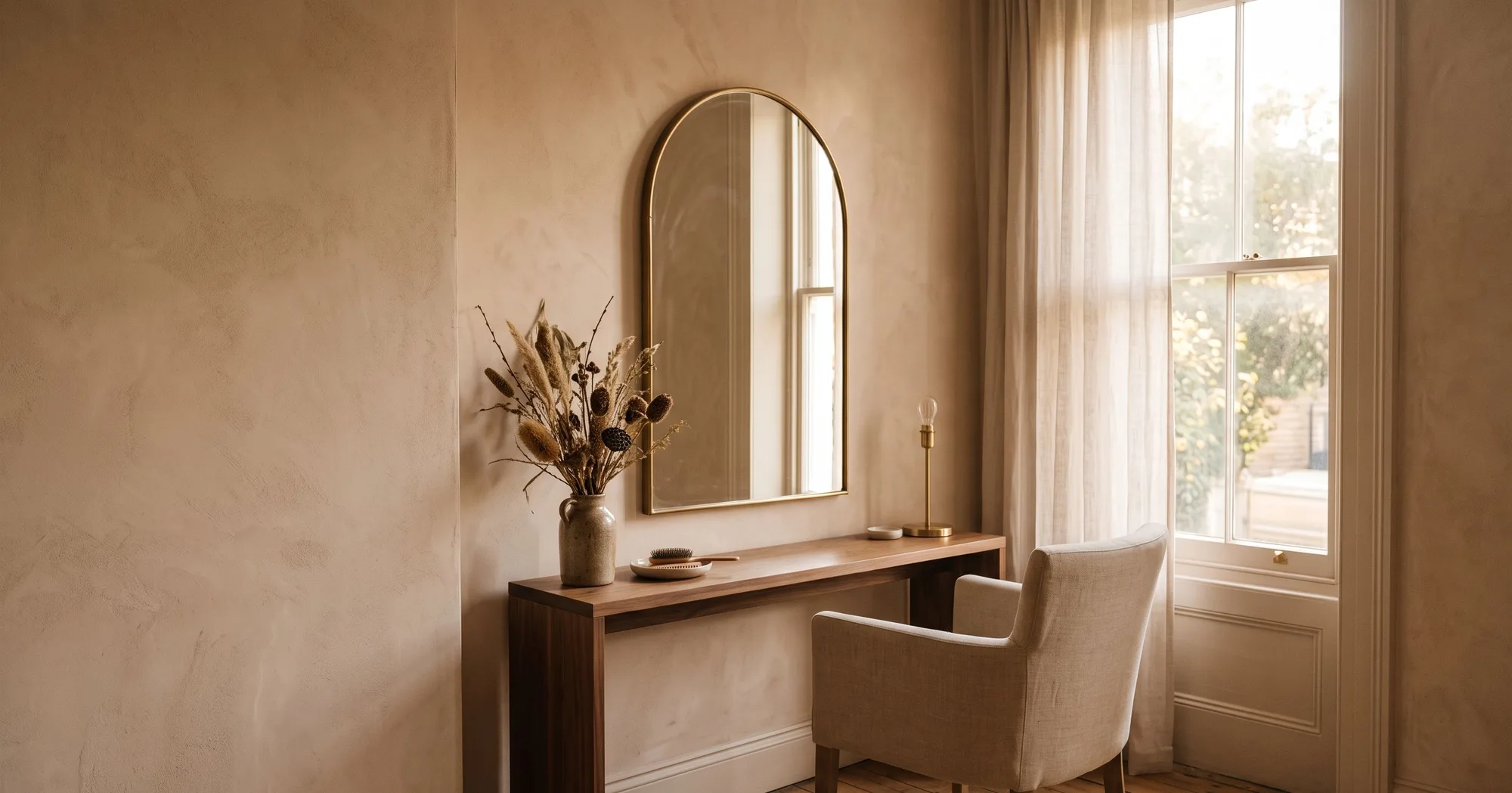 An arched mirror in a slim brass frame above a walnut ledge, against a warm plaster wall in late afternoon light.