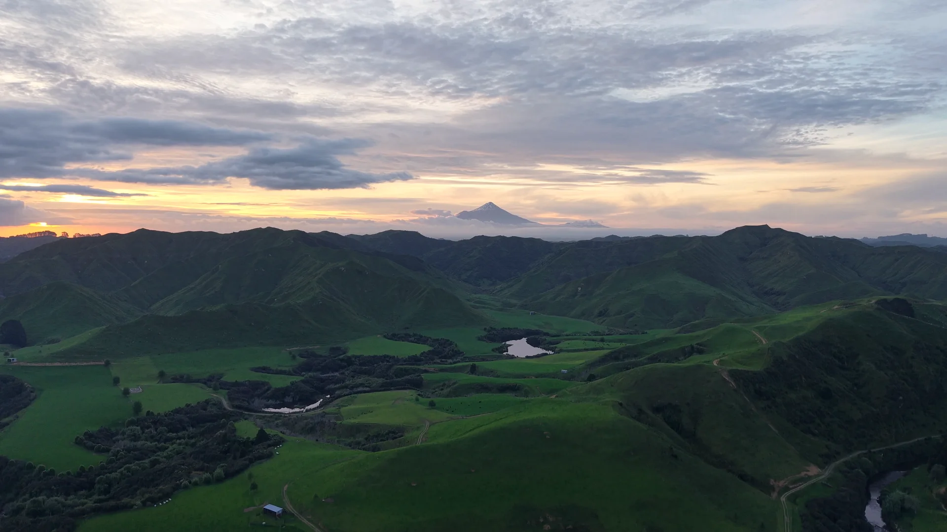 Aerial view of green New Zealand hill country at golden hour, with a volcanic peak on the horizon.