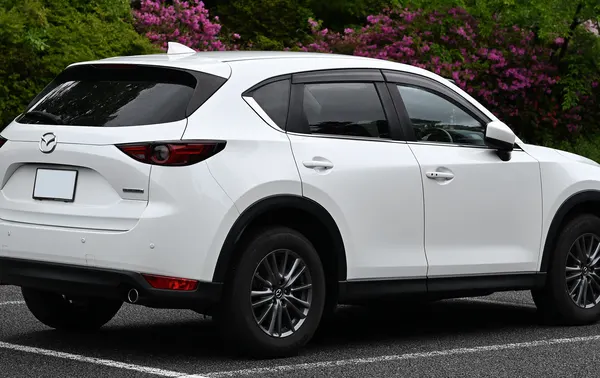 A white Mazda CX-5 SUV seen from the rear quarter in a car park with flowering shrubs behind.