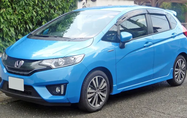 A bright blue Honda Fit hatchback parked on a street with trees behind it.